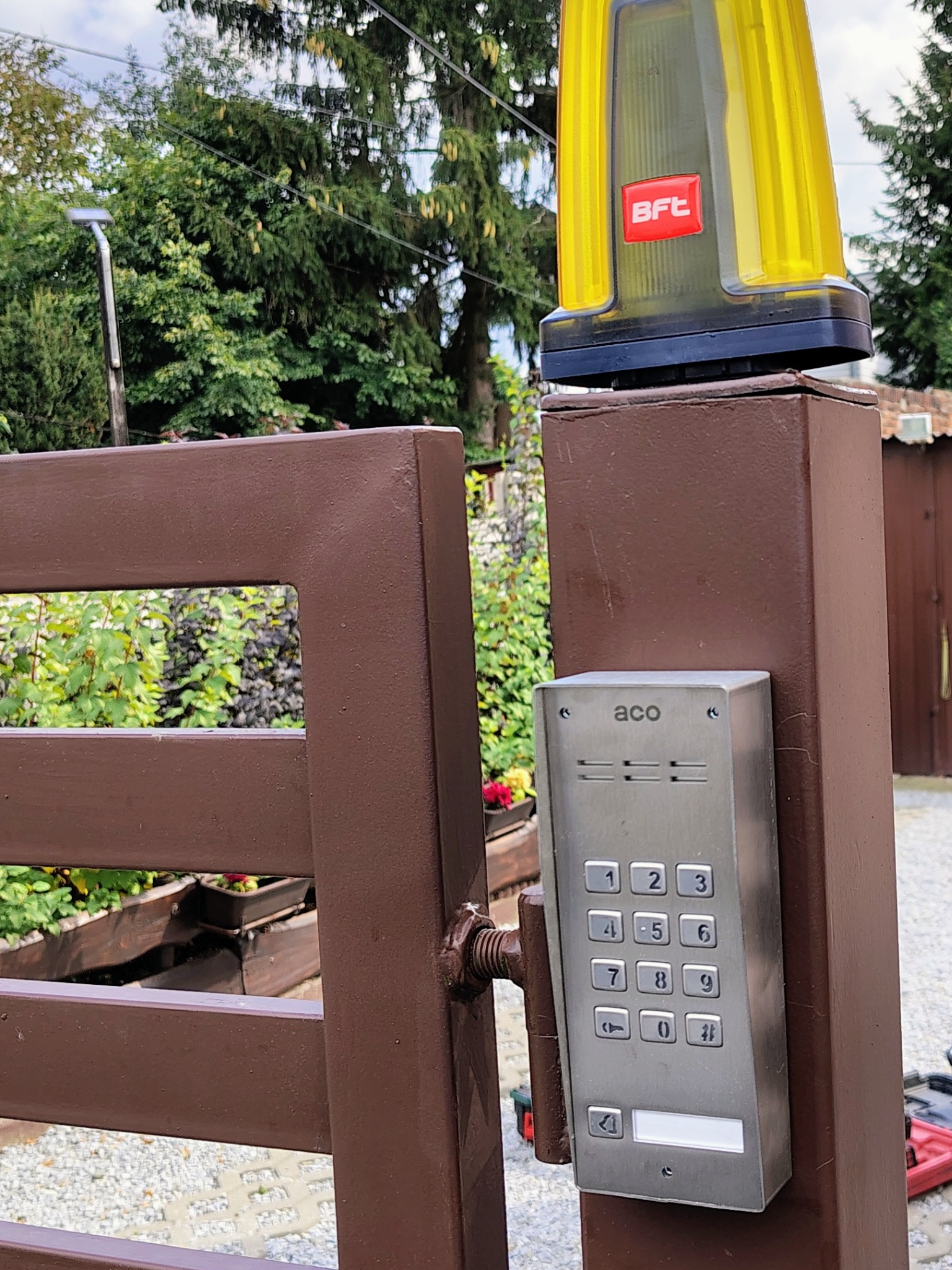 Brązowa brama z automatycznym systemem dostępu ACO, lampą ostrzegawczą BFT i widocznym ogrodem w tle.