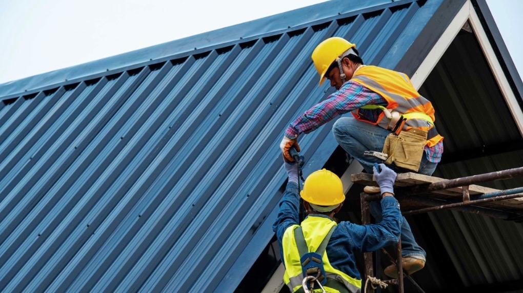 Dwóch dekarzy w kaskach i kamizelkach odblaskowych montuje blachę trapezową na dachu domu jednorodzinnego, widok z dołu na rusztowanie i fragment dachu.
