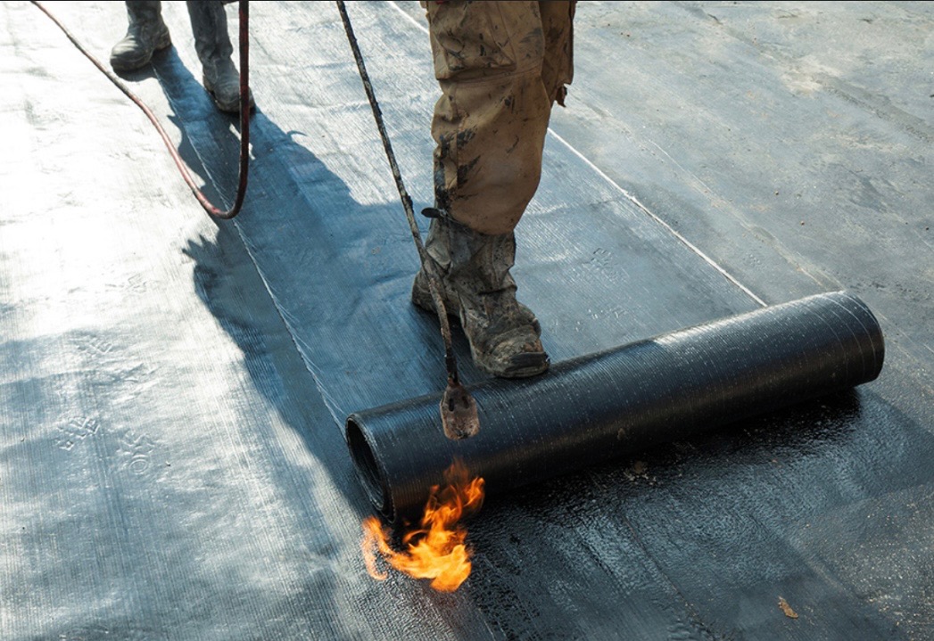 Dekarz w brudnych butach spawa papę termozgrzewalną na dachu, widoczny płomień palnika i rolka papy.