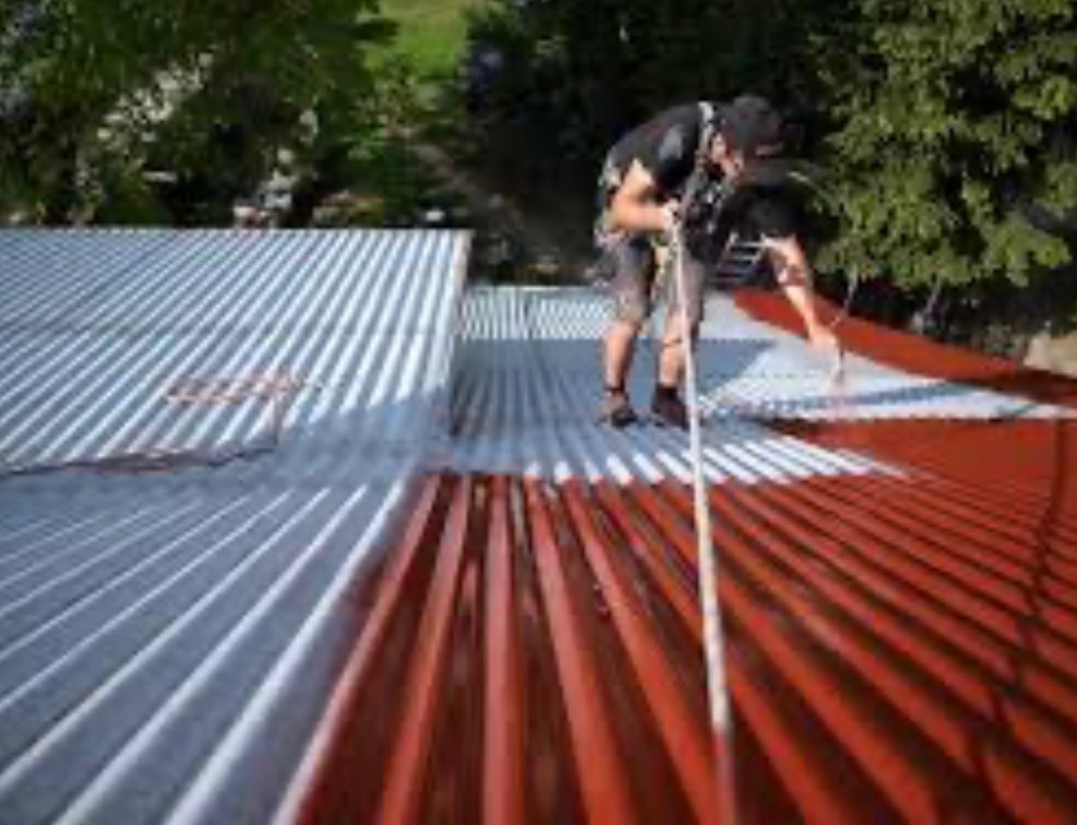 Pracownik w uprzęży maluje czerwoną farbą blachę trapezową na dachu, część dachu pomalowana na szaro.