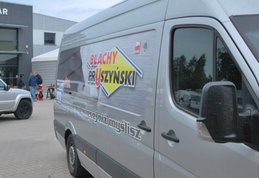 Srebrny samochód dostawczy z logo firmy Blachy Pruszyński na tle budynku firmy i zaparkowanego Jeepa Cherokee.