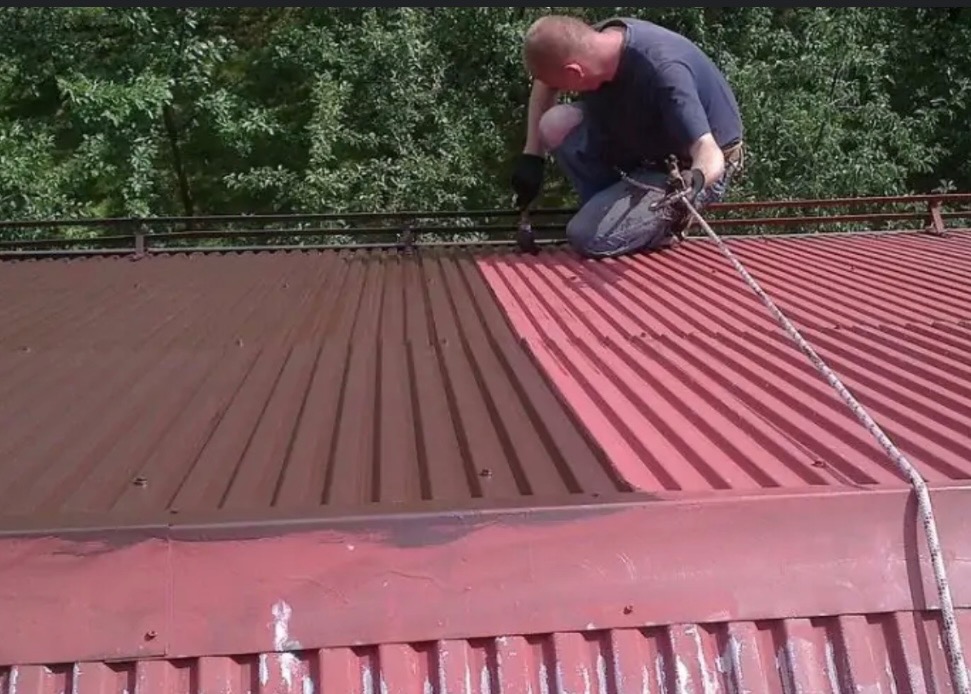 Pracownik w uprzęży maluje pędzlem dach z blachy trapezowej na brązowo, widoczna część dachu już pomalowana, druga w kolorze czerwonym, w tle drzewa.