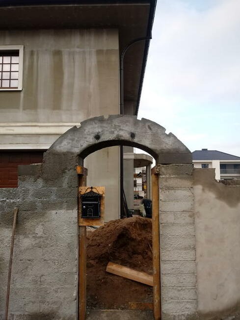 Wznoszona brama z łukowym zwieńczeniem z surowego betonu, widoczny mur z szarego betonu komórkowego, skrzynka pocztowa i pryzma ziemi wewnątrz budowanej bramy.