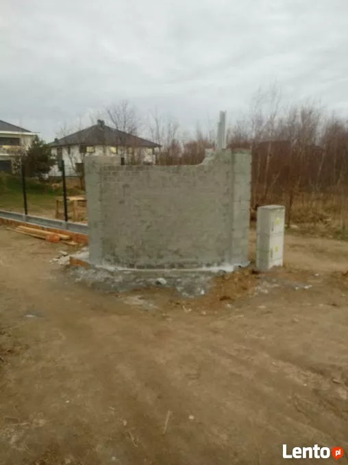 Wznoszenie betonowego muru na działce budowlanej, widoczne bloczki betonowe i zaprawa, w tle dom jednorodzinny i drzewa bezlistne.