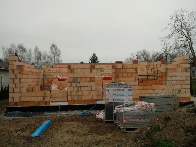 Wznoszone ściany domu z pustaków ceramicznych Porotherm, widoczne palety z materiałem budowlanym i niebieski element izolacyjny na ziemi.