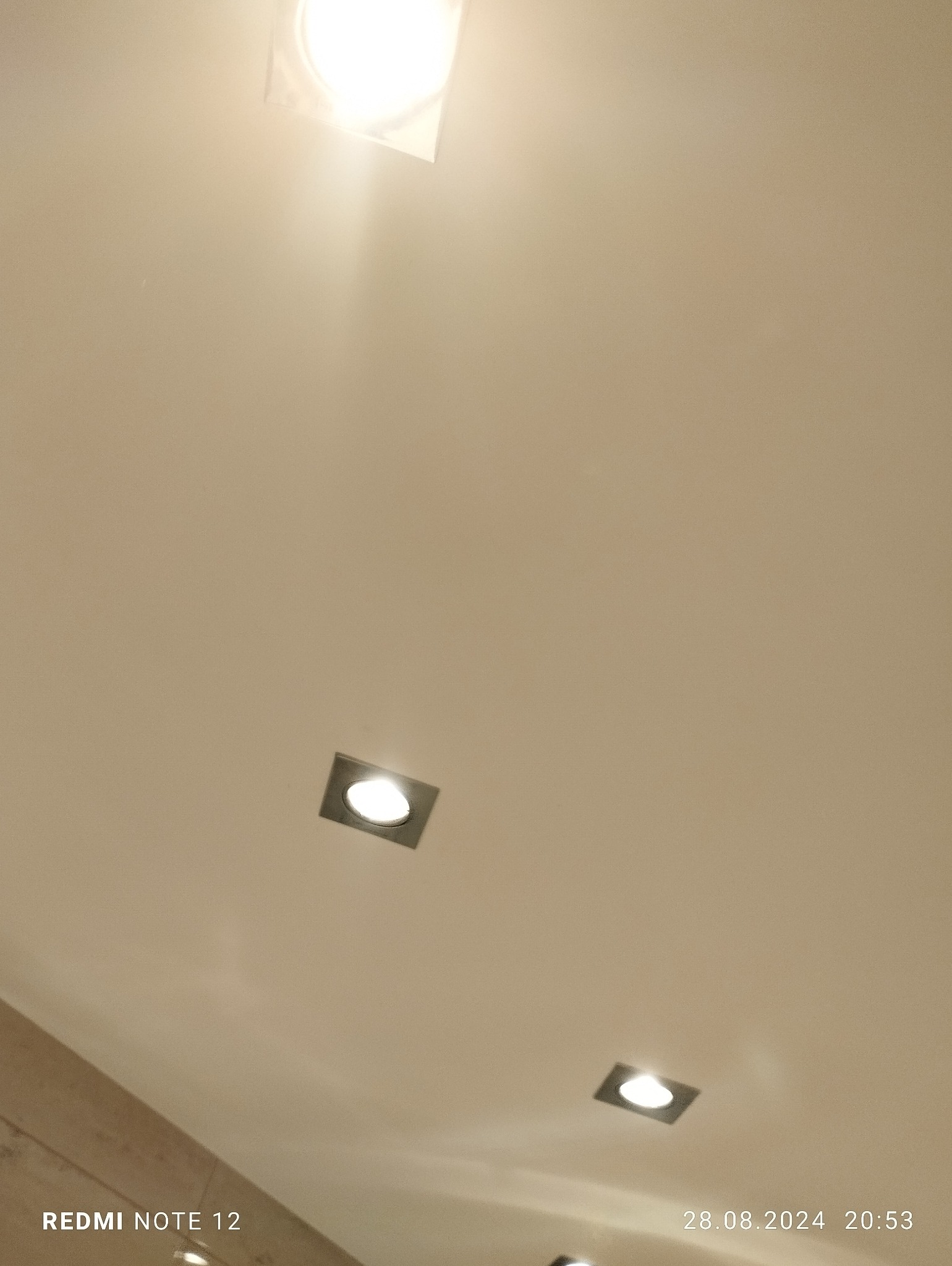 Biały sufit z trzema kwadratowymi lampami LED wpuszczanymi, widoczne z dołu pod kątem, z datą i modelem telefonu w rogu