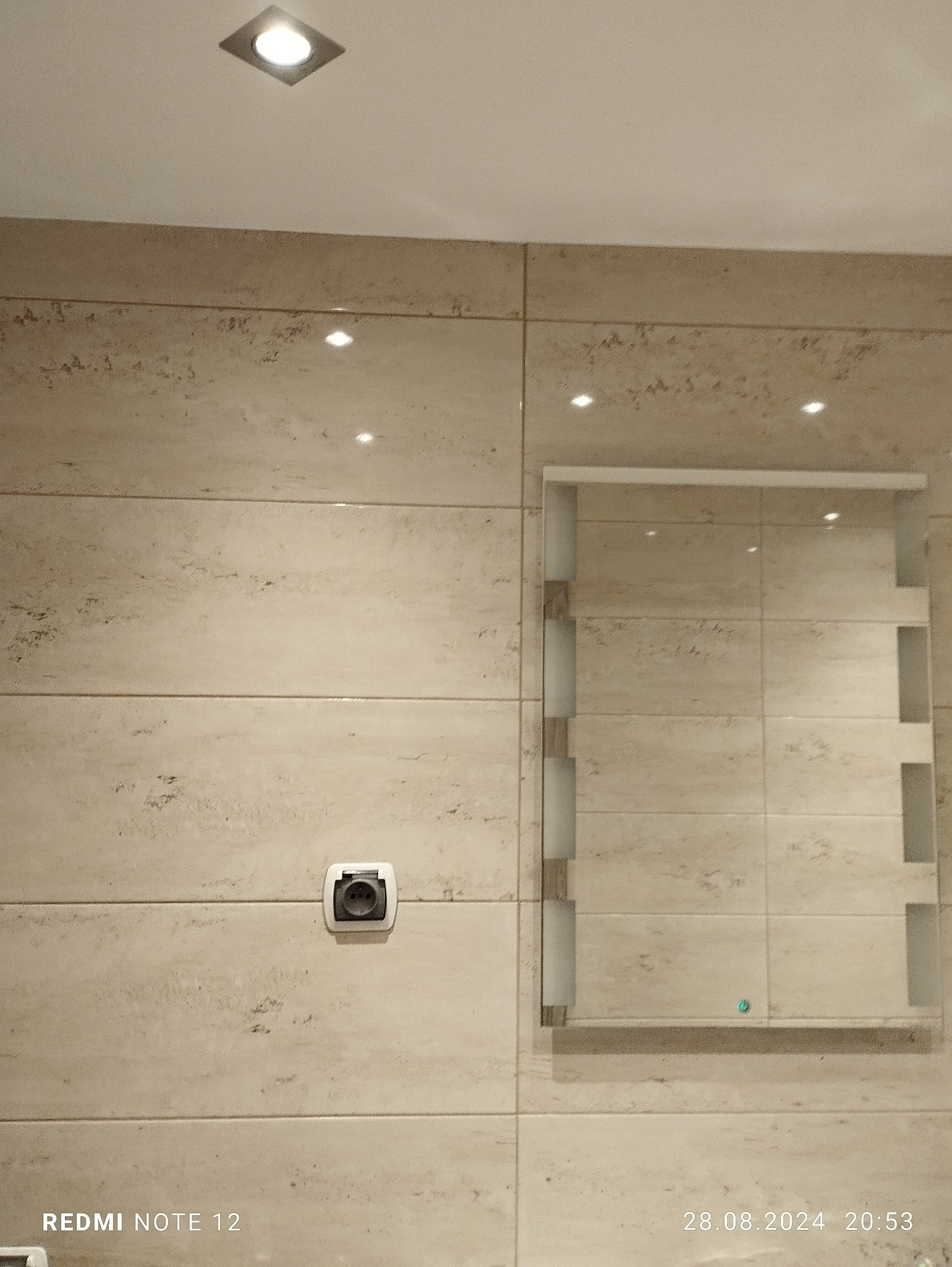 Wykończona ściana w łazience z beżowych, poziomych płytek imitujących kamień, z wbudowanym lustrem z podświetleniem bocznym i gniazdkiem elektrycznym z klapką ochronną.