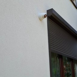 Wojciech Jarczewski - Kopułkowa kamera monitoringu CCTV zamontowana na elewacji budynku obok okna z brązową roletą zewnętrzną.