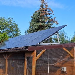 Wojciech Jarczewski - Panele słoneczne zamontowane na dachu drewnianej konstrukcji, otoczone zielenią i błękitnym niebem.