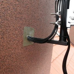 Wojciech Jarczewski - Kable w czarnej osłonie przymocowane do ściany budynku o chropowatej, brązowej fakturze, zabezpieczone szarą masą uszczelniającą przy wejściu do budynku. Widoczny fragment dachu z dachówką.