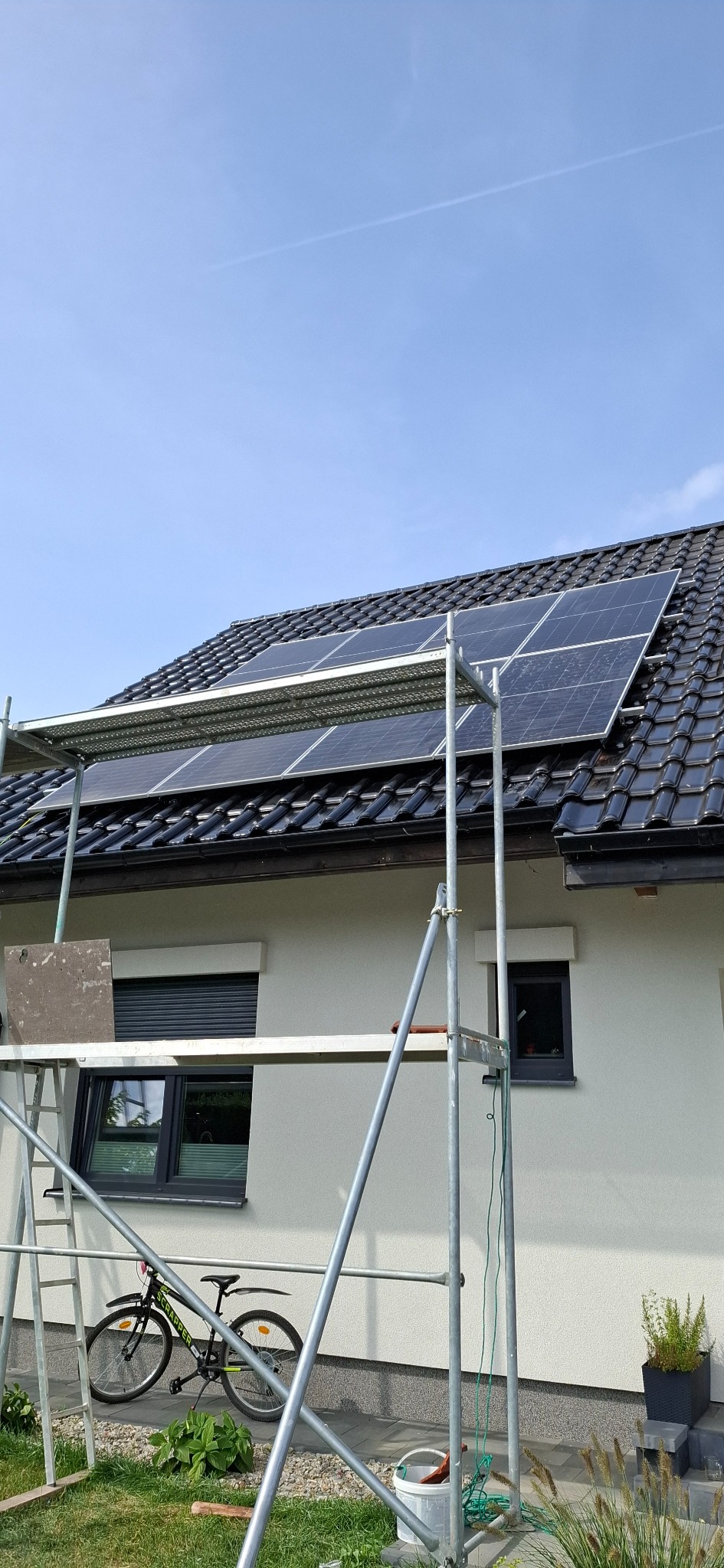 Montaż paneli słonecznych na dachu domu jednorodzinnego, widoczne rusztowanie, rower oparty o ścianę, błękitne niebo w tle.