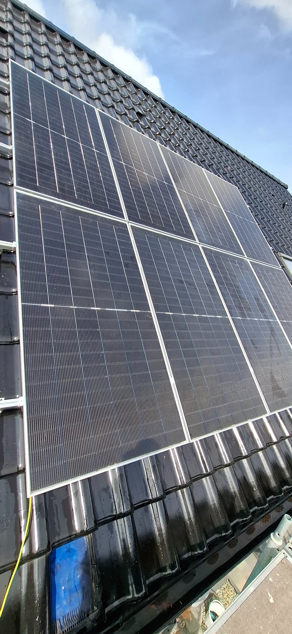 Panele fotowoltaiczne zamontowane na błyszczącym, czarnym dachu z dachówki karpiówki, widoczne narzędzia i niebieska plandeka zabezpieczająca.