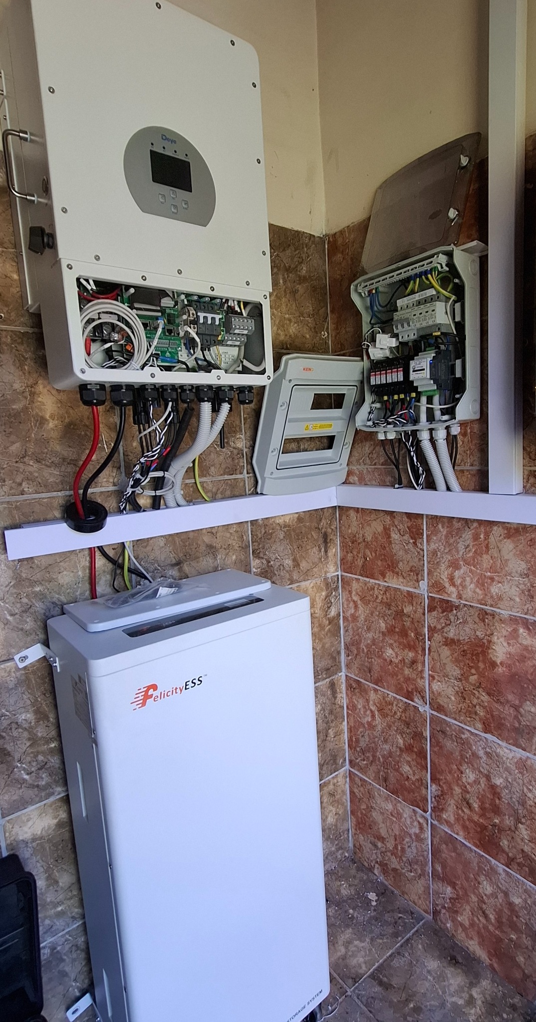 Biały falownik z otwartą obudową, biała skrzynka rozdzielcza z otwartymi drzwiczkami i biały magazyn energii Felicity ESS, zamontowane na ścianie wyłożonej brązowymi płytkami.