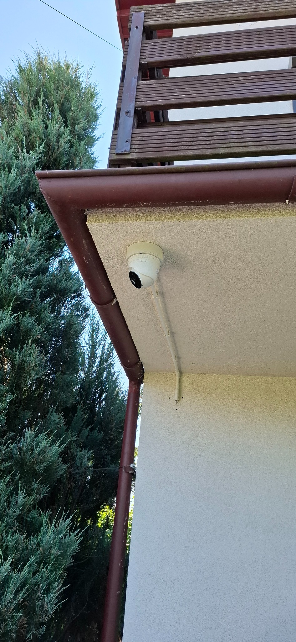 Kamera monitoringu marki HiLook zamontowana pod okapem domu, obok rynny i iglastego drzewa. Widoczne przewody instalacyjne.