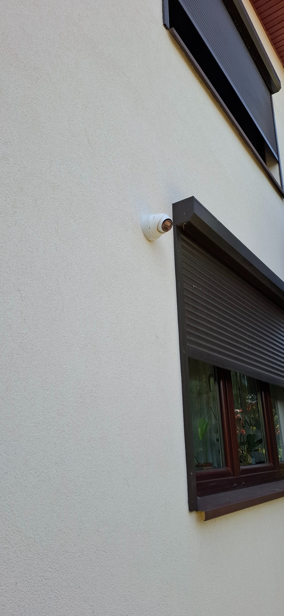 Kopułkowa kamera monitoringu CCTV zamontowana na elewacji budynku obok okna z brązową roletą zewnętrzną.
