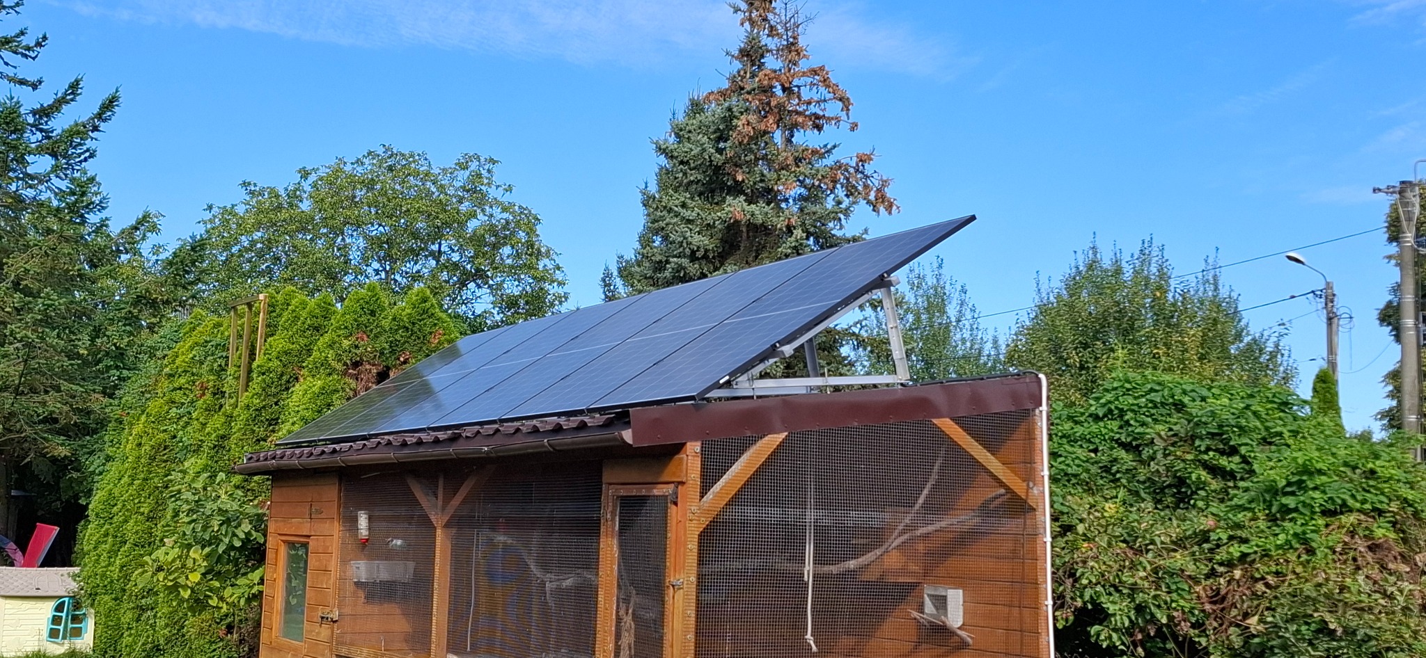 Panele słoneczne zamontowane na dachu drewnianej konstrukcji, otoczone zielenią i błękitnym niebem.