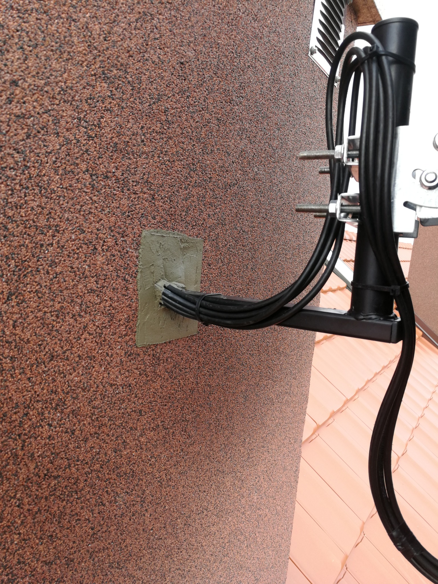 Kable w czarnej osłonie przymocowane do ściany budynku o chropowatej, brązowej fakturze, zabezpieczone szarą masą uszczelniającą przy wejściu do budynku. Widoczny fragment dachu z dachówką.