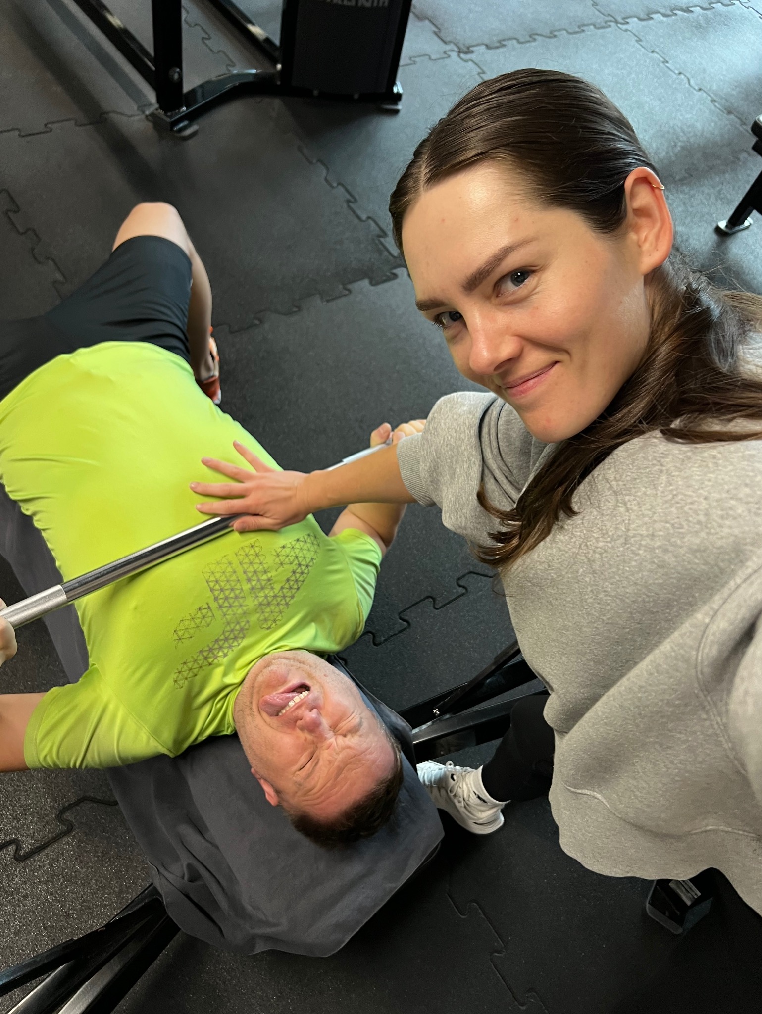 Mężczyzna w neonowej koszulce ćwiczy wyciskanie sztangi leżąc, a kobieta w szarej bluzie asekuruje go i uśmiecha się do kamery.