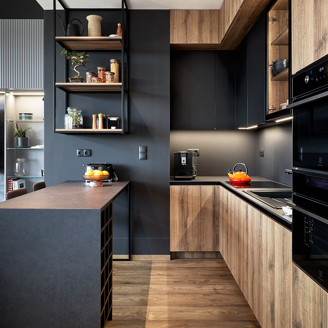 Nowoczesna kuchnia z drewnianymi frontami szafek i czarnymi elementami, oświetlenie podszafkowe, wyspa kuchenna z blatem w kolorze grafitu, półki wiszące z dekoracjami, widoczny ekspres do kawy...