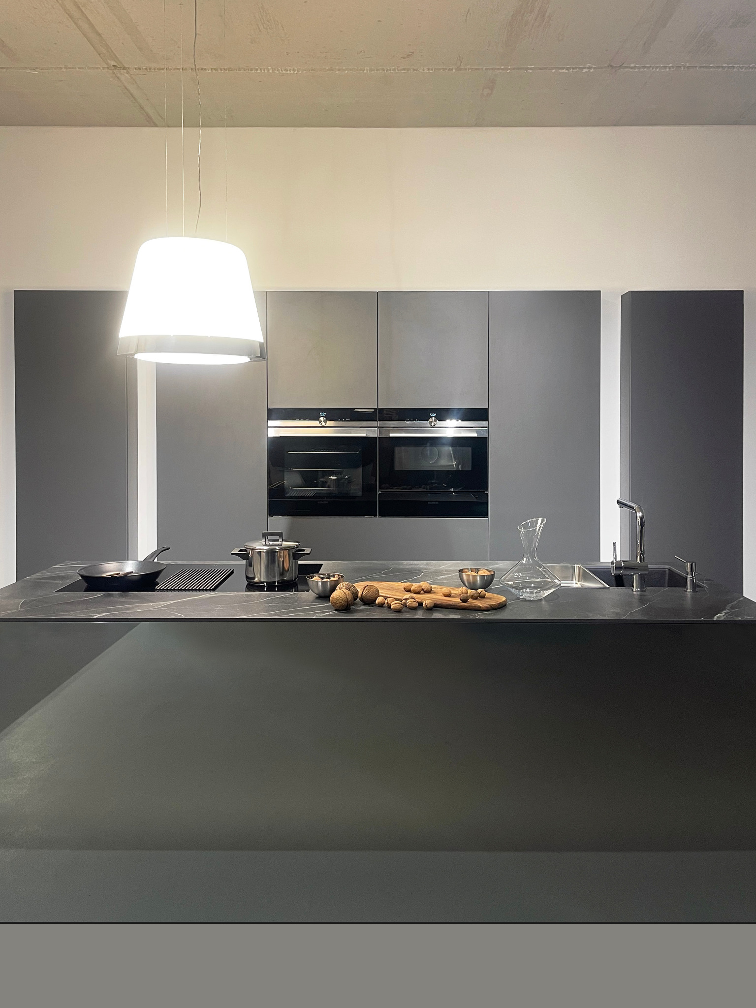 Nowoczesna kuchnia w odcieniach szarości z kamiennym blatem, na którym znajdują się naczynia, owoce i karafka, oświetlona wiszącą lampą.