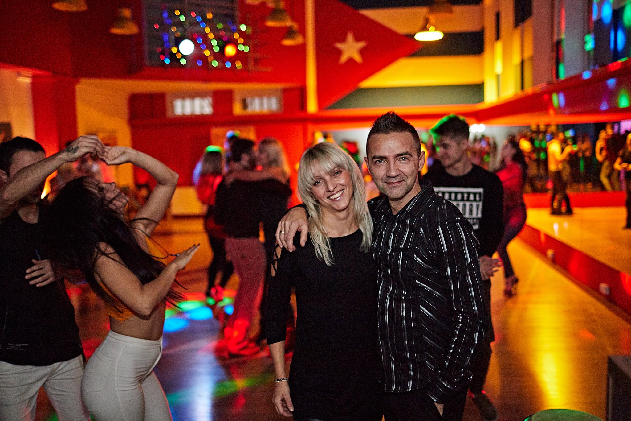 Para uśmiechnięta, obejmująca się na tle sali tanecznej z flagą Kuby i tańczącymi ludźmi w tle.