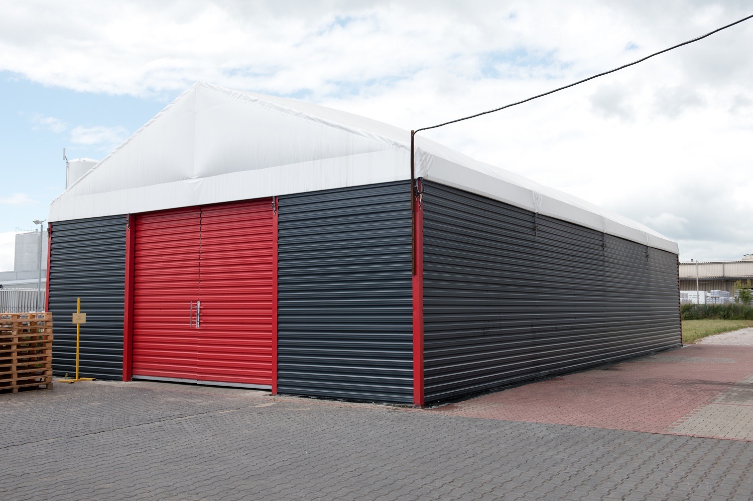 Gotowa hala magazynowa z szarej blachy trapezowej z czerwonymi drzwiami rolowanymi, biały dach, widoczne palety i fragment placu z kostki brukowej.