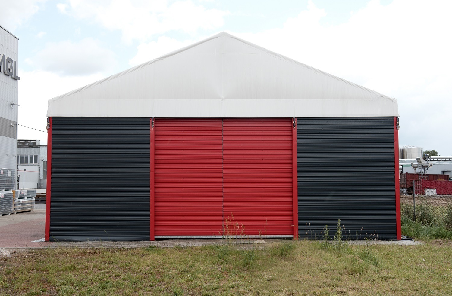 Gotowa hala magazynowa z czerwonymi drzwiami rolowanymi i szarą elewacją, widok z zewnątrz na tle zachmurzonego nieba.