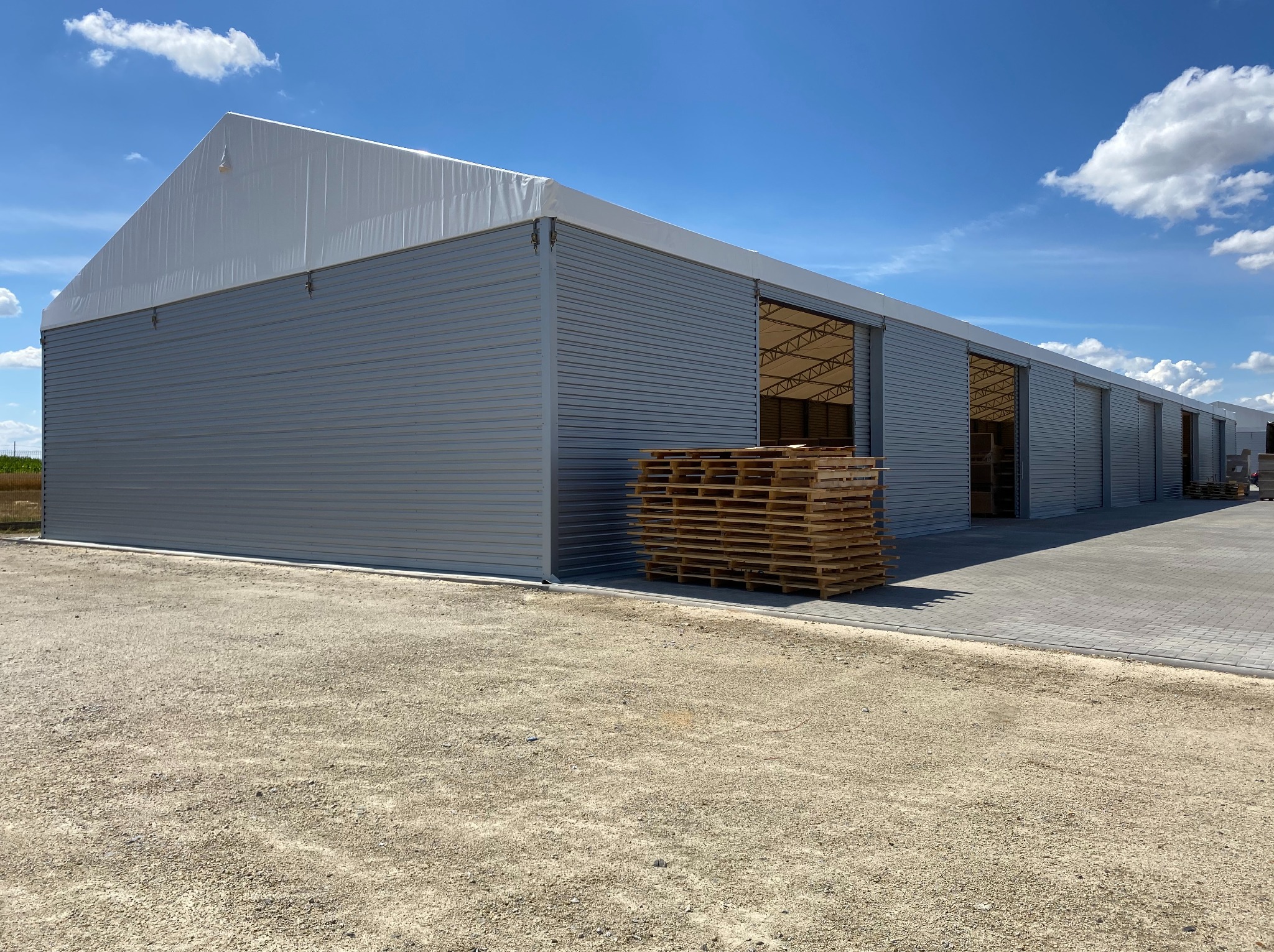 Długa hala magazynowa z szarej blachy falistej i białym dachem namiotowym, z otwartymi wrotami i stertą palet przed nią, na tle błękitnego nieba z chmurami.
