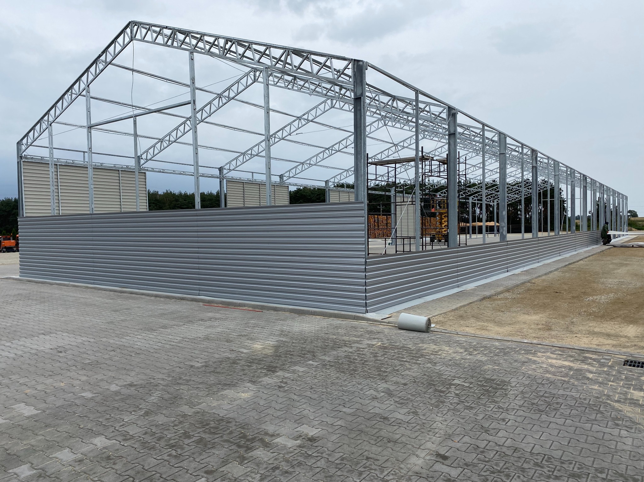 Stalowa konstrukcja hali w trakcie montażu, widoczne szare panele ścienne, rusztowania i częściowo utwardzony teren.