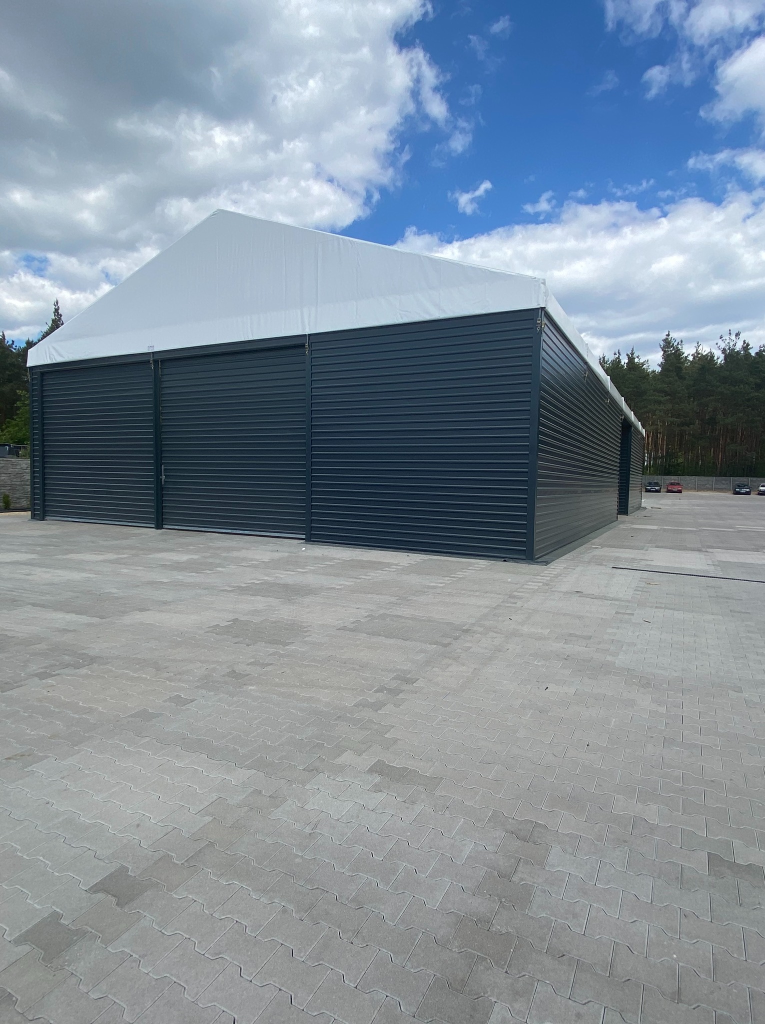 Gotowa hala z blachy trapezowej w kolorze grafitowym z białym dachem membranowym, widoczne przesuwne bramy wjazdowe, na zewnątrz wyłożona kostką brukową.