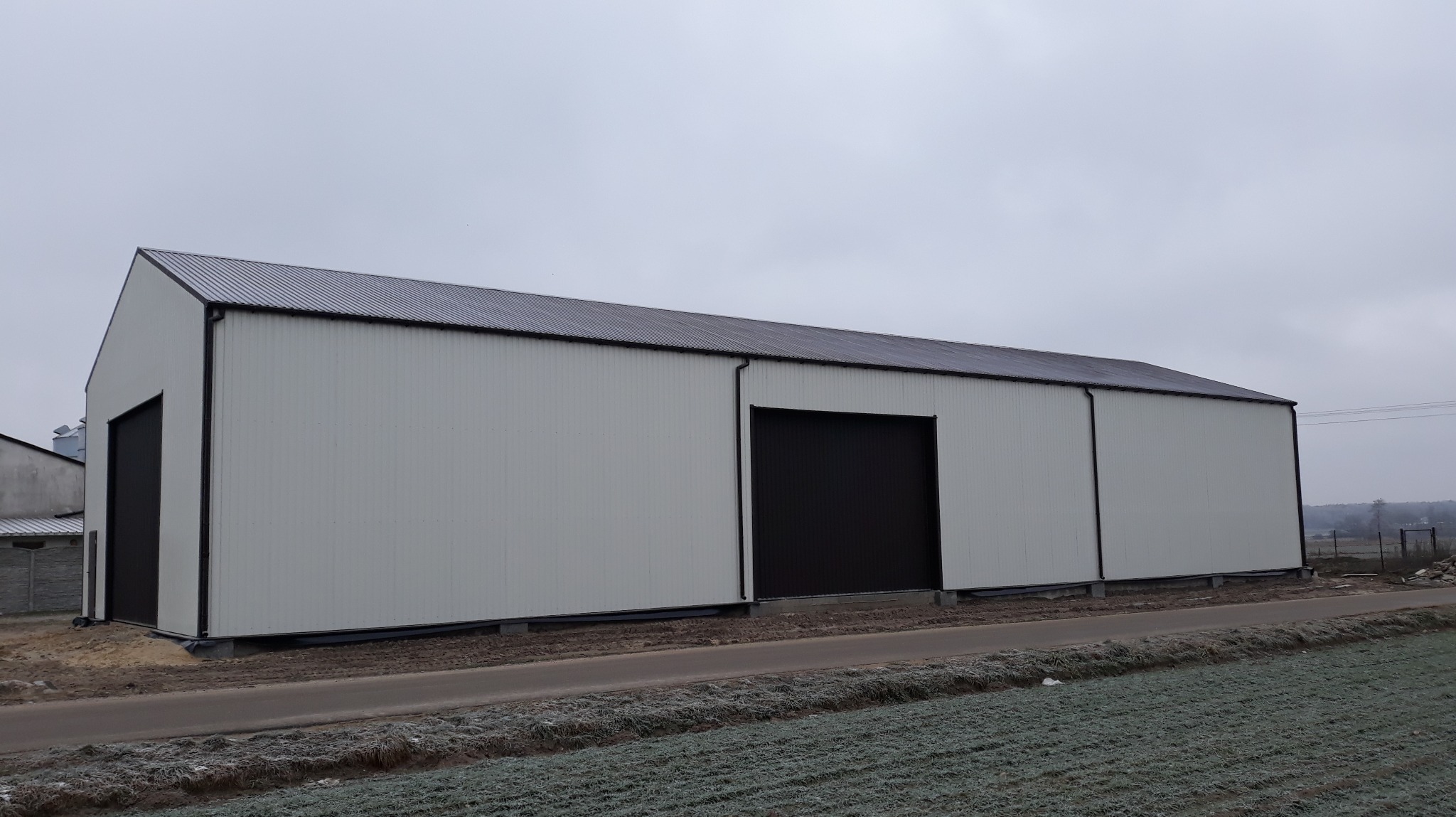 Zewnętrzny widok dużej, nowej hali z białymi ścianami i ciemnymi, przesuwnymi bramami, stojącej na obrzeżach pola w pochmurny dzień.