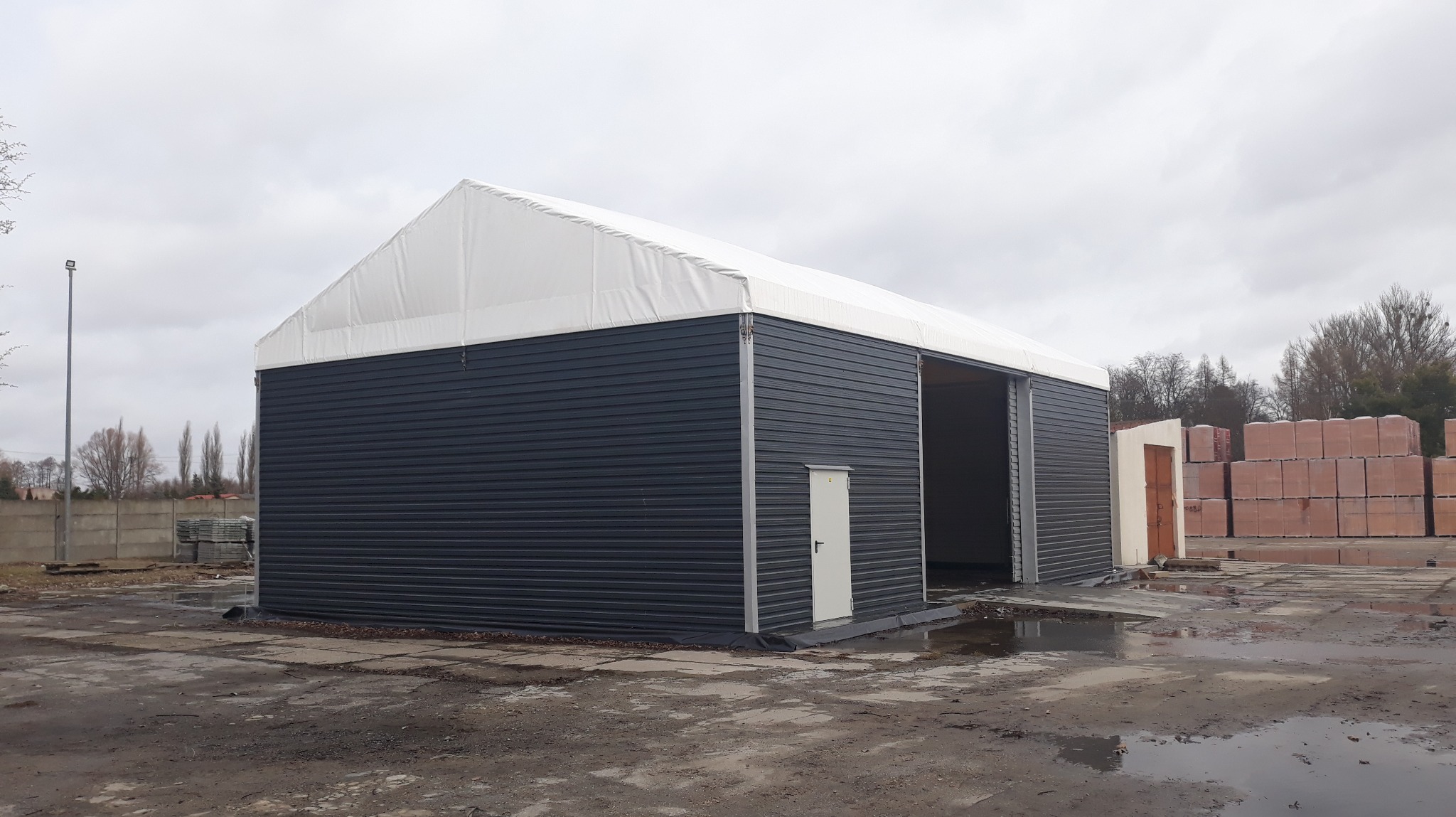 Gotowa hala magazynowa z szarej blachy trapezowej i białym dachem membranowym, widoczna boczna brama wjazdowa i drzwi wejściowe, obok składowane palety z cegłami.