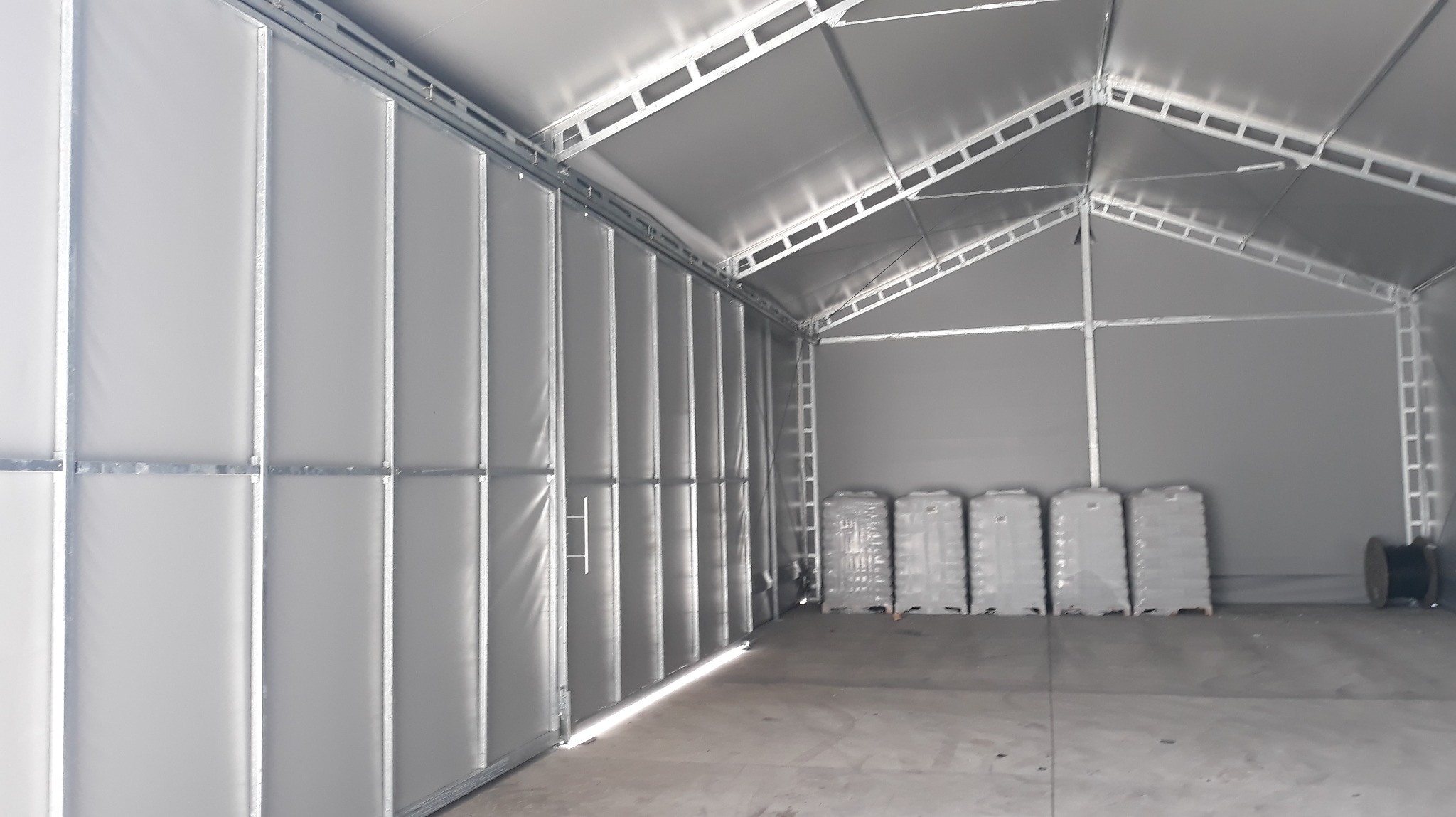 Wnętrze nowej hali z metalową konstrukcją, szarymi ścianami z paneli i paletami w tle, widok z perspektywy wejścia.