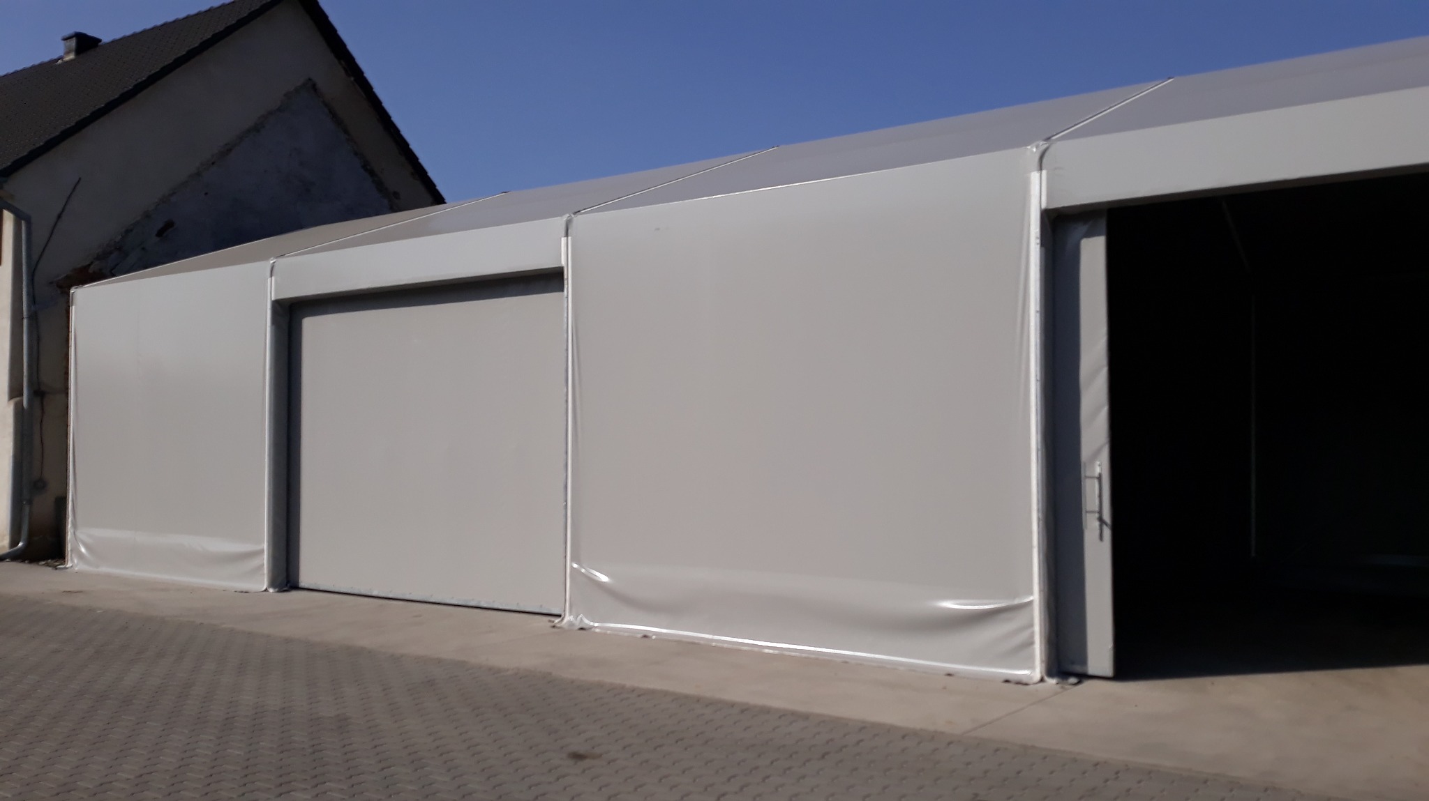 Szara hala namiotowa z przesuwnymi drzwiami i otwartym wjazdem, widoczny fragment budynku mieszkalnego w tle, jasne niebo.