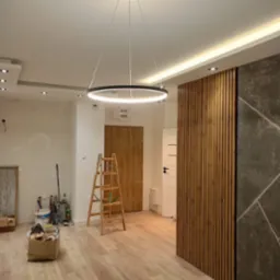 Wnętrze w trakcie remontu z zamontowanym okrągłym, nowoczesnym oświetleniem LED, drewnianą ścianką dekoracyjną, widoczna drabina i narzędzia malarskie.