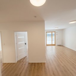 ProStyle - Jasne, przestronne wnętrze po remoncie: drewnopodobne panele, białe ściany, drzwi z przeszkleniami i nowoczesne oświetlenie sufitowe. Widok na balkon.