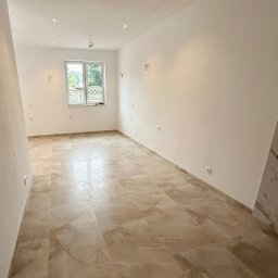 ProStyle - Jasne wnętrze po remoncie: beżowe płytki podłogowe z marmurkowym wzorem, białe ściany z widocznymi gniazdkami i oknem. Minimalistyczny design, gotowe do umeblowania.