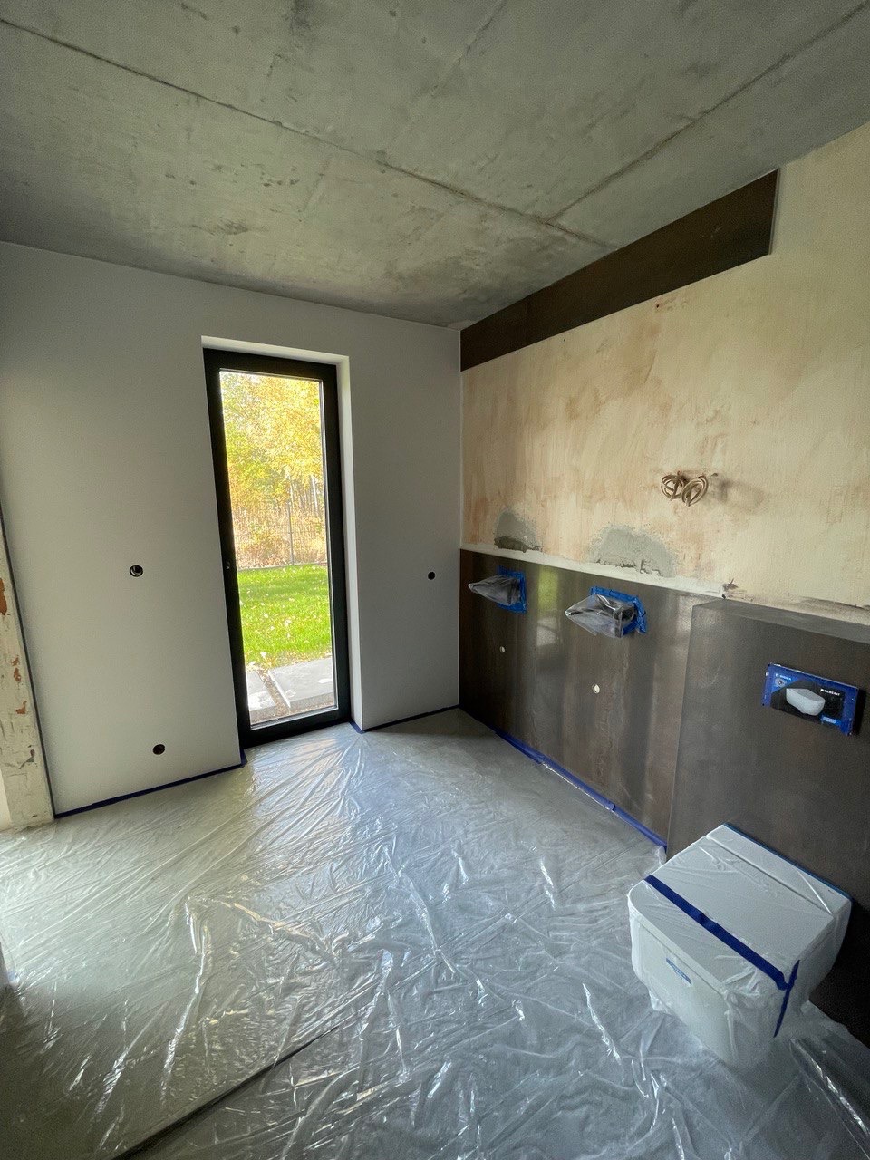 Przygotowanie do remontu łazienki: zabezpieczona folią podłoga, częściowo wykończone ściany z zamontowanymi elementami instalacji, widoczna surowa betonowa płyta stropowa.