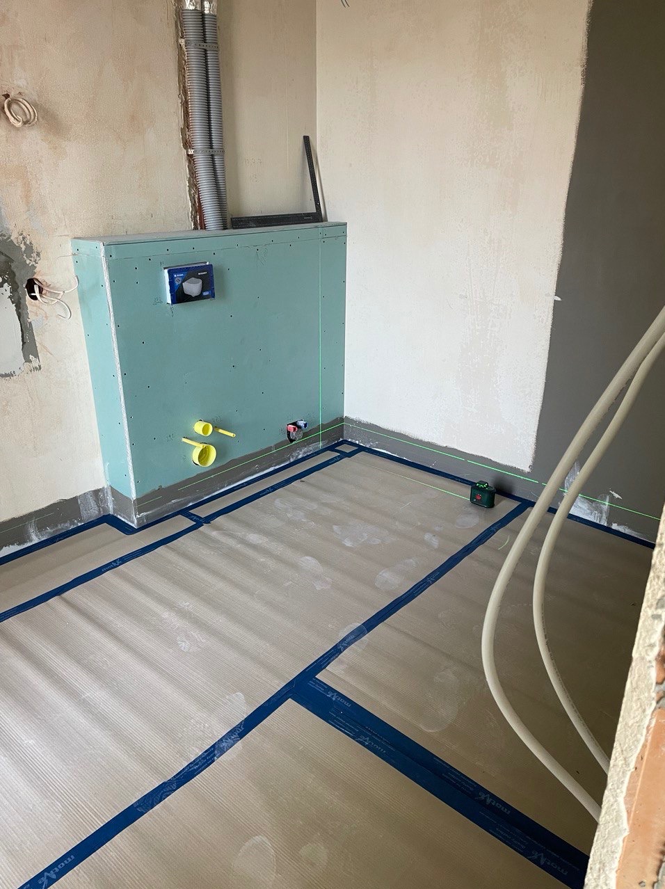 Przygotowanie podłoża pod płytki w łazience: mata izolacyjna z niebieską taśmą, zabudowa stelaża WC z instalacją hydrauliczną, poziomica laserowa.