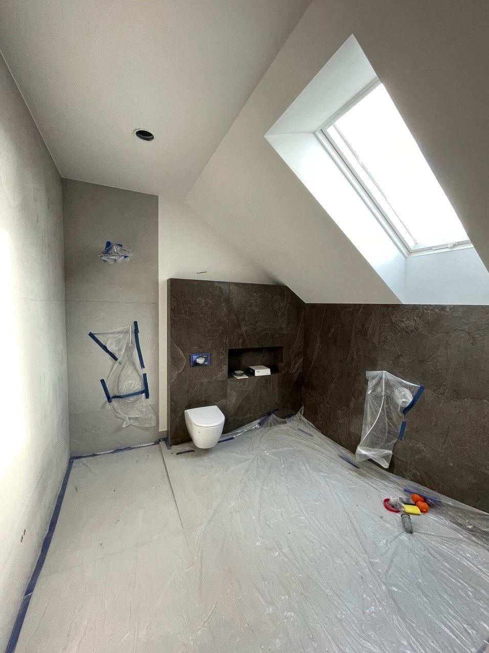 Łazienka w trakcie prac wykończeniowych: szare płytki imitujące kamień, podwieszana toaleta, okno dachowe, folia ochronna na podłodze, zabezpieczona strefa prysznica.