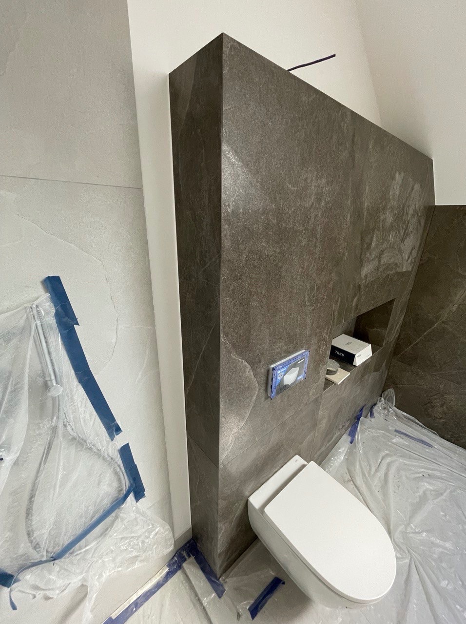 Montaż podwieszanej toalety z szarą okładziną ścienną imitującą kamień, zabezpieczona folią ochronną podczas prac wykończeniowych w łazience.