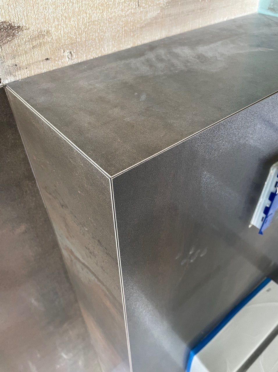 Detale narożnika obudowy z płytek imitujących metal, z widocznymi listwami wykończeniowymi, na tle ściany z widoczną strukturą.