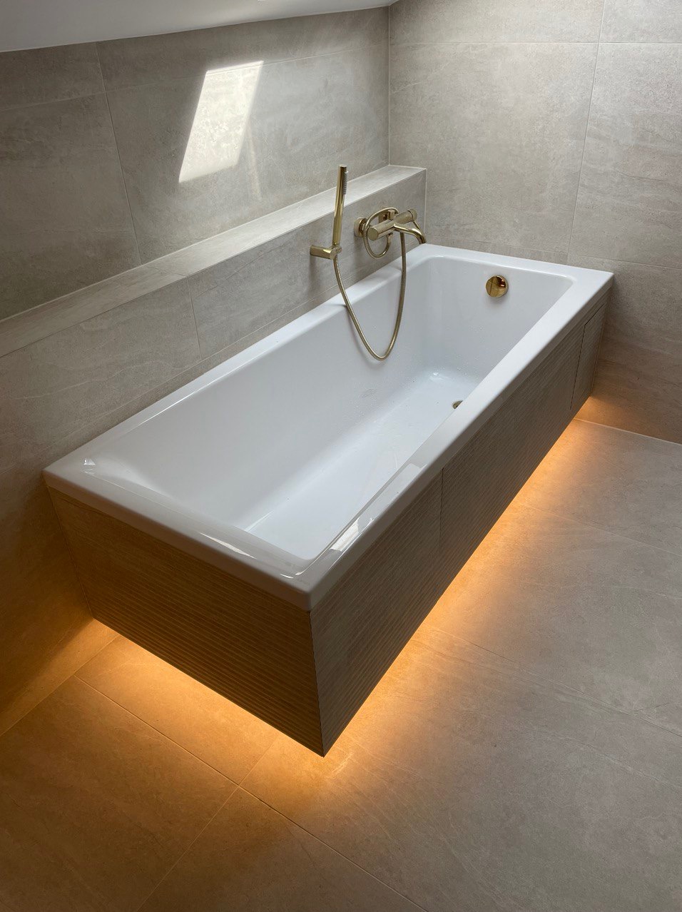 Biała wanna obudowana płytkami imitującymi drewno, z podświetleniem LED od dołu, złota bateria, jasne płytki na ścianach i podłodze, światło dzienne wpadające z okna dachowego.