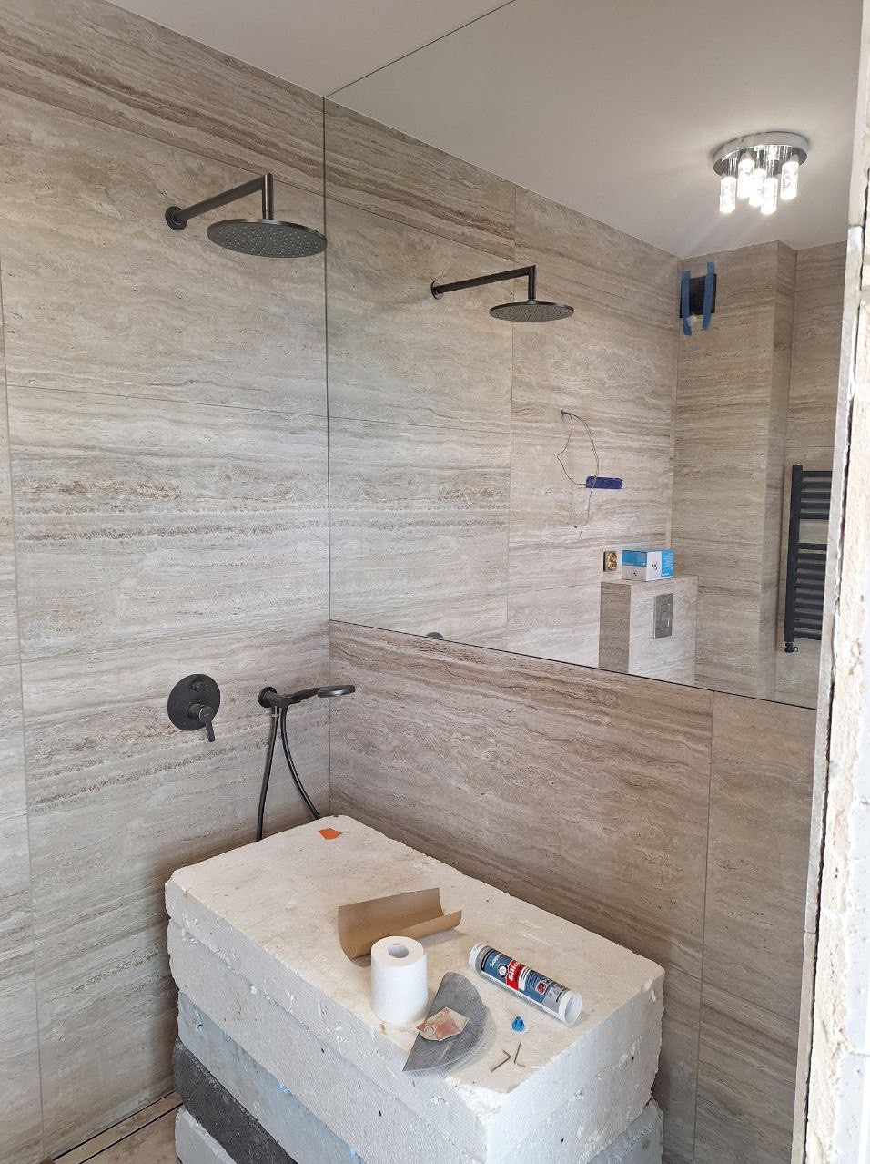 Montaż armatury w łazience w Łomiankach: widok na podwójny prysznic z deszczownicą, baterią podtynkową i ułożone płytki imitujące trawertyn. Na pierwszym planie zaplecze glazurnicze.