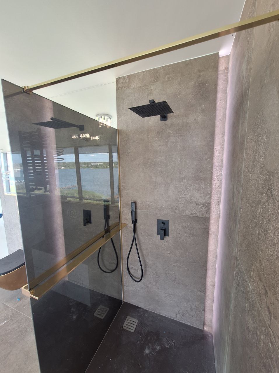 Nowoczesna kabina prysznicowa walk-in z czarną armaturą, szklaną ścianką z złotymi profilami i widokiem na jezioro za oknem.