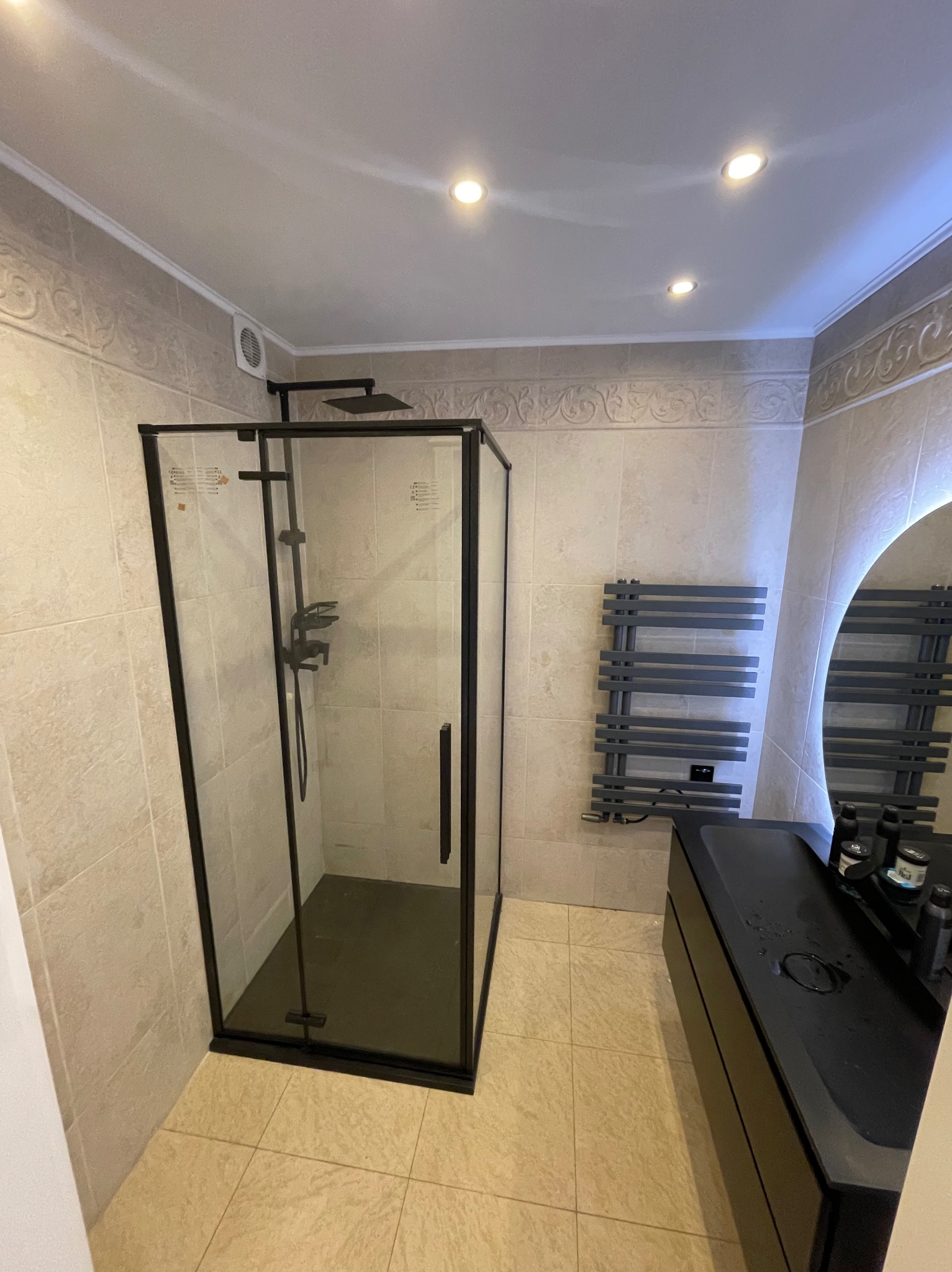 Nowoczesna łazienka z kabiną prysznicową typu walk-in z czarnymi profilami, grzejnikiem drabinkowym i czarną umywalką nablatową z okrągłym lustrem podświetlanym LED.