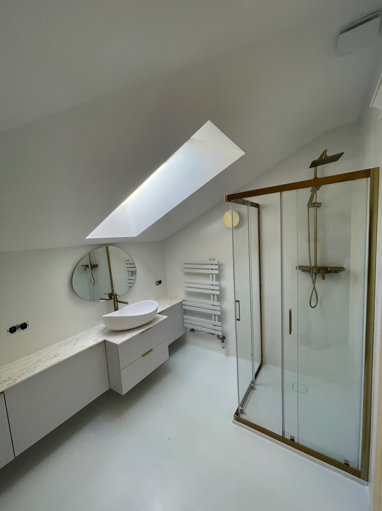 Nowoczesna łazienka na poddaszu z oknem dachowym, białą armaturą, kabiną prysznicową z elementami w kolorze złotym, owalną umywalką na szafce z marmurowym blatem i grzejnikiem drabinkowym.