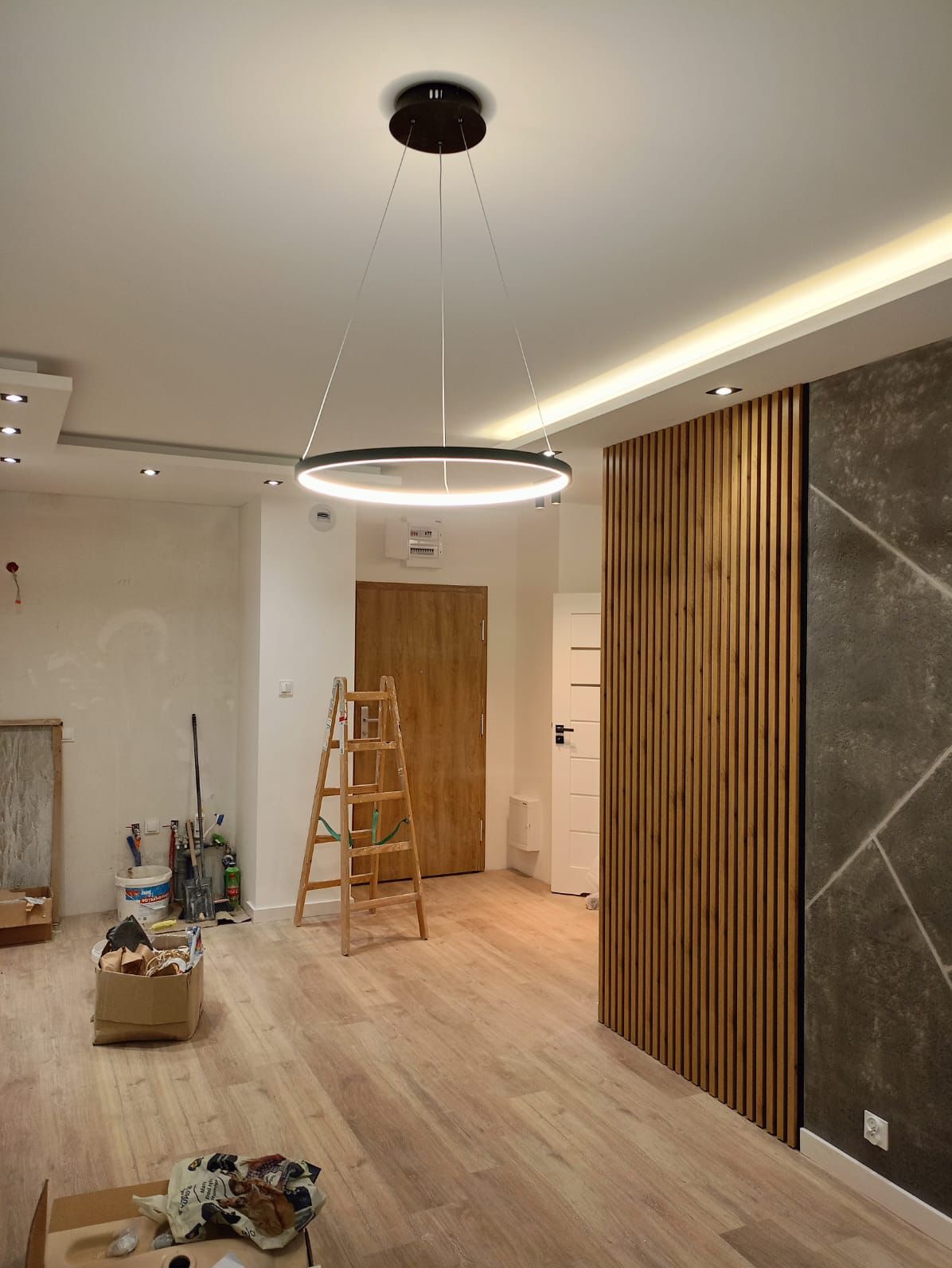 Wnętrze w trakcie remontu z zamontowanym okrągłym, nowoczesnym oświetleniem LED, drewnianą ścianką dekoracyjną, widoczna drabina i narzędzia malarskie.