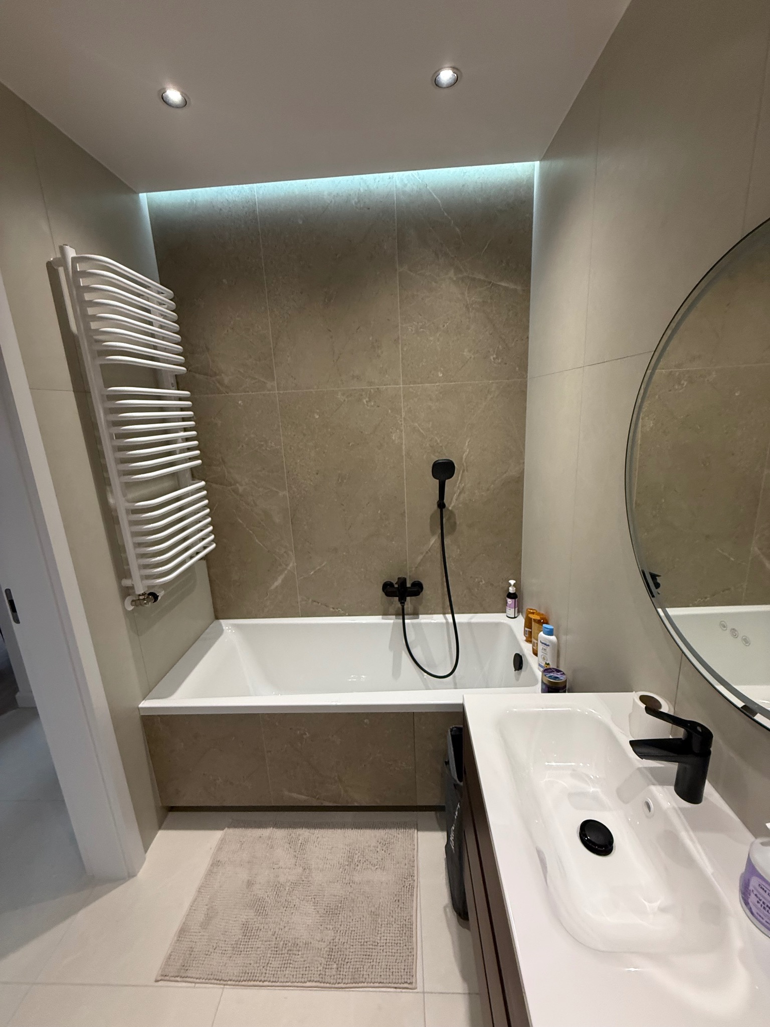 Nowoczesna łazienka z beżowymi płytkami, białą wanną i umywalką z czarną armaturą. Grzejnik drabinkowy i okrągłe lustro dopełniają wystrój.