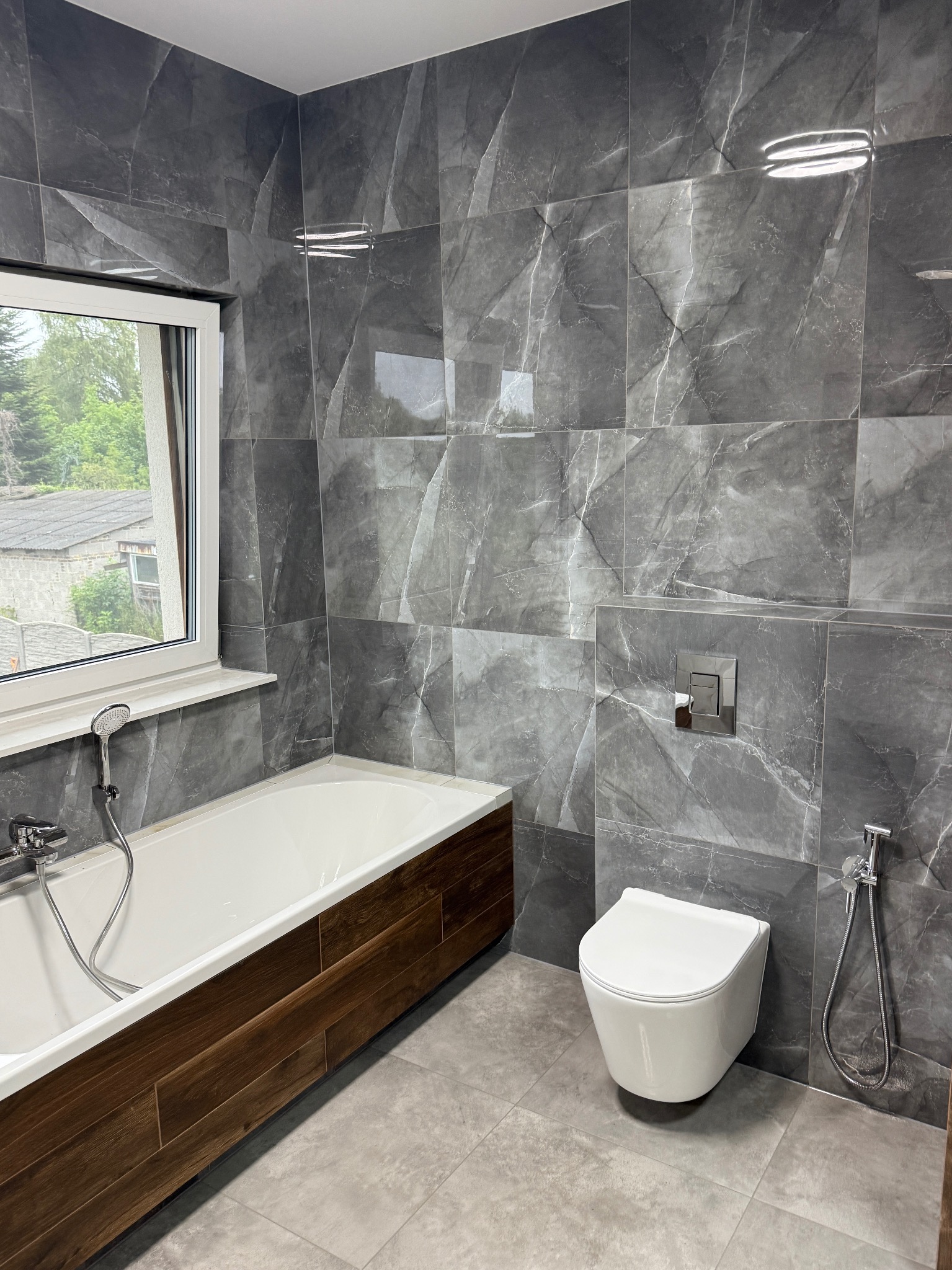 Wykończona łazienka z szarymi płytkami imitującymi marmur, białą wiszącą toaletą i wanną z drewnianą obudową. Widok na zewnątrz przez okno.